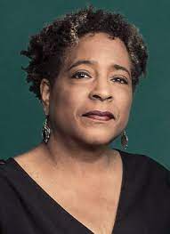 Deborah Gray White, a Black woman with short, curly hair, is pictured with brown lipstick. She wears a black shirt in front of a green background. 